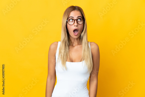 Young blonde woman isolated on yellow background looking up and with surprised expression