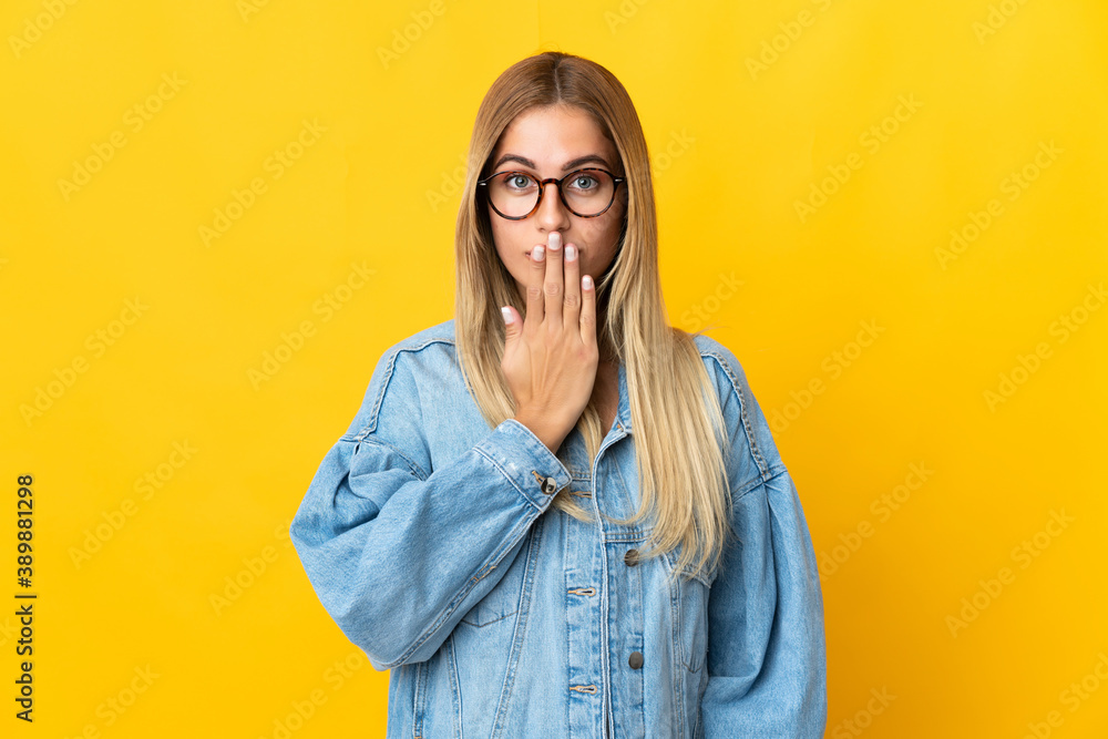 Young blonde woman isolated on yellow background covering mouth with hand