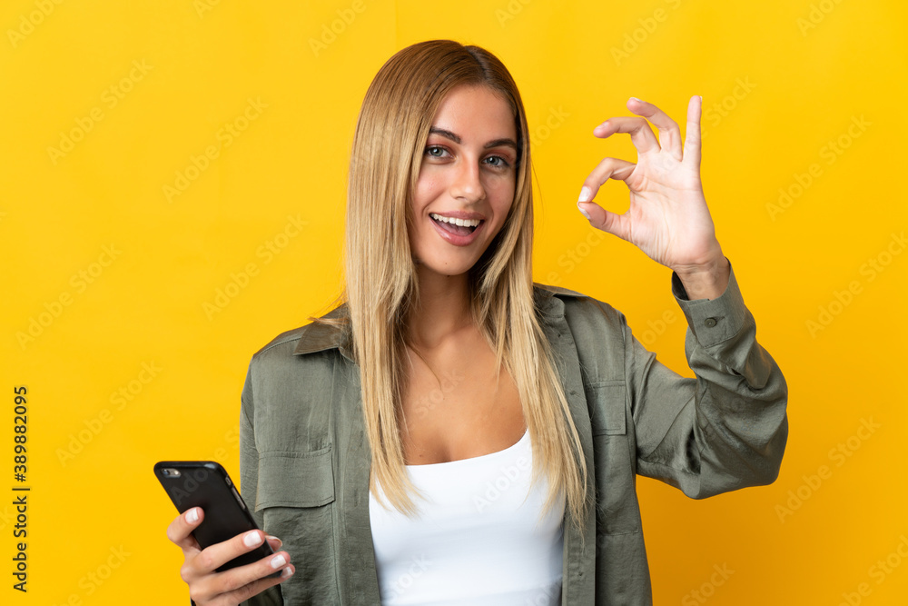 Young blonde woman isolated on yellow background using mobile phone and doing OK sign Young blonde woman isolated on yellow background using mobile phone and doing OK sign