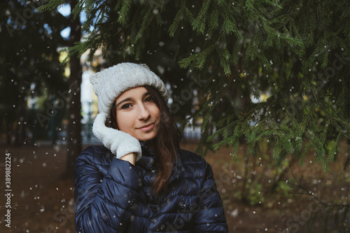 .Portrait of a happy girl in a knitted hat, gloves and a red Christmas sweater among a winter fir forest and falling snow. The concept of the first snow, Christmas holidays and winter fashion.