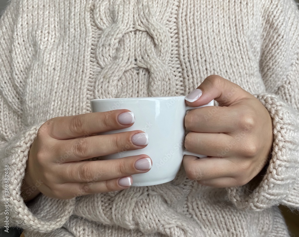 Beautiful French manicure. Well-groomed female hands with painted nails hold a white coffee mug on a cozy beige knitted woolen sweater background. Fashionable color of nail gel polish. Beautiful French manicure. Well-groomed female hands with painted nails hold a white coffee mug on a cozy beige knitted woolen sweater background. Fashionable color of nail gel polish.