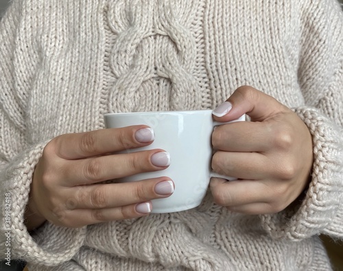 Beautiful French manicure. Well-groomed female hands with painted nails hold a white coffee mug on a cozy beige knitted woolen sweater background. Fashionable color of nail gel polish.