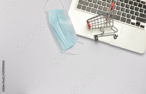 Shopping trolley and medical face mask with laptop on white background. Covid-19 pandemic. Online shopping. Selective focus.