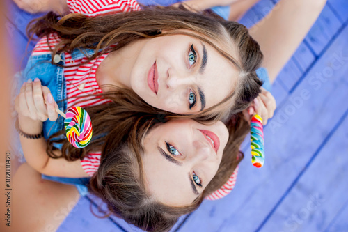 Top view of portrait twin sisters in colorful clothes with lollipops sitting on violet boards