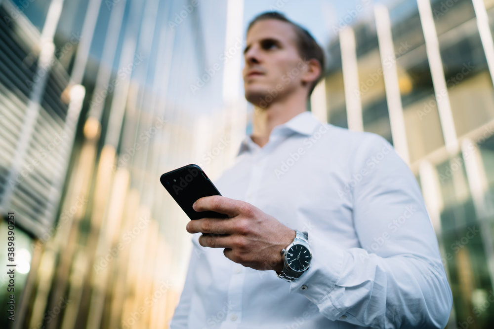 Below view on blurred Caucasian businessman with wearable swiss watch on hand holding modern smartphone device and thinking in financial district, male entrepreneur using mobile gadget in downtown Below view on blurred Caucasian businessman with wearable swiss watch on hand holding modern smartphone device and thinking in financial district, male entrepreneur using mobile gadget in downtown