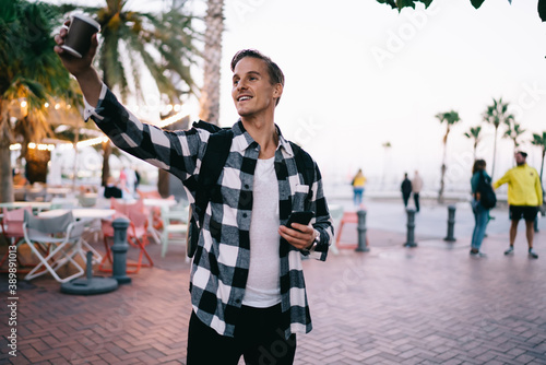 Smiling caucasian hipster guy in casual trendy wear and with backpack gesture raising hand with coffee to go cup on street, positive male male traveler enjoying exploring city on free time vacations #389891013