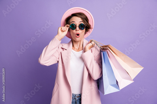 Photo of amazed pretty optimistic bob hair young lady hold bags wear glasses pink cap coat isolated on purple color background