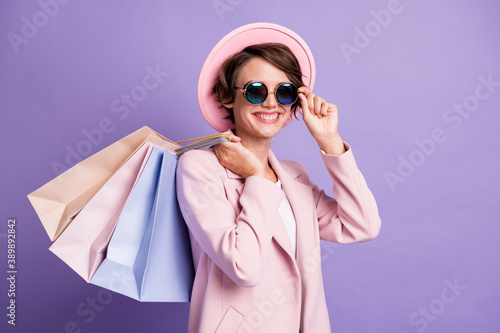 Portrait of adorable sweet brunette hair young lady hold bags touch glasses wear pink cap coat isolated on purple color background