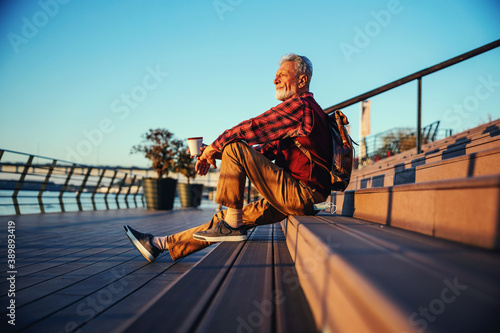 Side view of bearded hipster senior man sitting on the stairs outdoors, drinking coffee and looking at river. photo