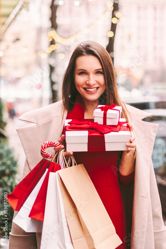 Cheerful young attractive woman hold gift boxes and paper shopping bags in hands, smile on city street. Beautiful happy girl shopaholic with Christmas gifts after holiday shopping in mall. Winter sale