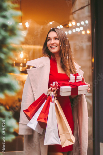 Young happy attractive woman hold shopping bags and gift boxes in hands and smile on city street. Beautiful cheerful girl with Christmas presents after holiday sale shopping in mall. Lady shopper