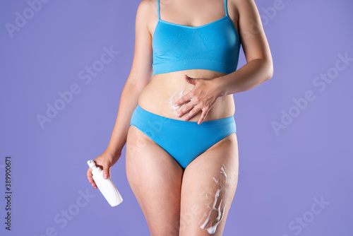Slim woman in blue underwear applying anti-cellulite mousse on purple background