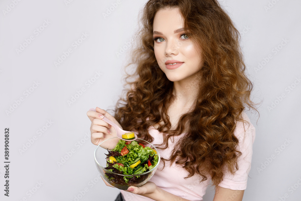 Curly girl on gray background. Portrait of beautiful smiling healthy woman holding a fresh vegetable salad bowl. Curly girl on gray background. Portrait of beautiful smiling healthy woman holding a fresh vegetable salad bowl.