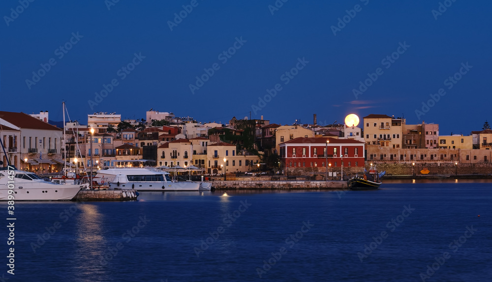 Full moon setting over Chania's Old Venetian Harbour. Night view of piers, boats and Maritime museum of Crete and houses over marina.