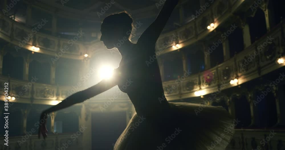 Cinematic shot of an young graceful classical ballet female dancer in white tutu is performing a choreography on classic theatre stage with dramatic lighting before start of a show.