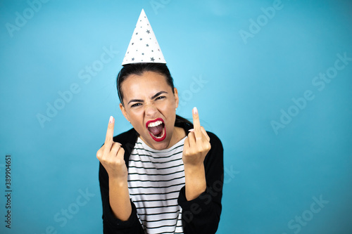 Obraz na płótnie Young beautiful woman wearing a birthday hat over insolated blue background showing middle finger doing fuck you bad expression, provocation and rude attitude