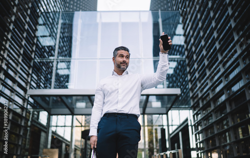 Confident businessman dressed in formal white shirt holding modern cellphone waving while see office partner at street, successful boss with smartphone gadget raising hand near corporate company