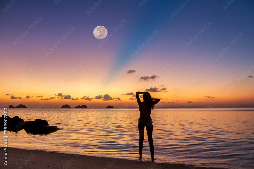 Silhouette of a young woman standing on the sandy beach by the sea against the backdrop of a spectacular sunset and a huge rising full moon on a summer evening