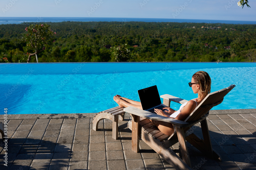 Female tourist using laptop for browsing while resting near swimming pool