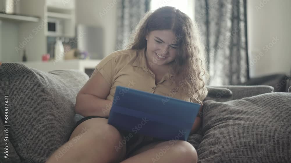 Portrait of cheerful plump woman using social media on laptop and smiling. Young overweight Caucasian lady sitting on couch and surfing Internet indoors. Lifestyle and wireless communication. Portrait of cheerful plump woman using social media on laptop and smiling. Young overweight Caucasian lady sitting on couch and surfing Internet indoors. Lifestyle and wireless communication.