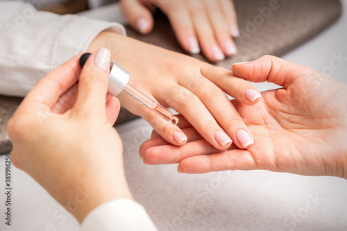 Hand of manicurist pours oil by pipette to cuticle of nails of young woman in beauty salon. French manicure