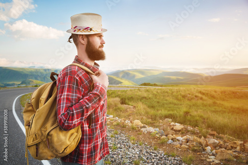 Young male tourist hitchhiker in a hat shorts with a backpack walks along a country asphalt road against the backdrop of mountains at sunset
