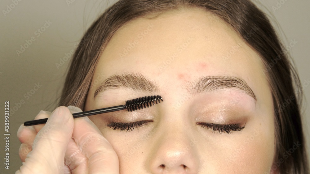 Master combs the eyebrows with a brush. Beautiful attractive female face of a brunette well-groomed woman or lady. Eyebrow lamination and styling. Stylist's hands. Permanent makeup for eyebrows. 