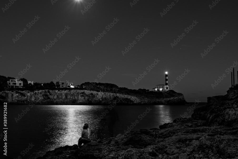 young woman sitting in front of ocean with moonlight reflection and lighthouse at night. poetic and relaxing image, mindfulness, in porto colom