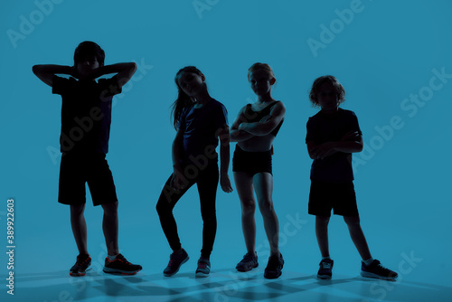Silhouetted full length shot of four little sportive kids looking at camera while posing, standing isolated over blue background © Svitlana