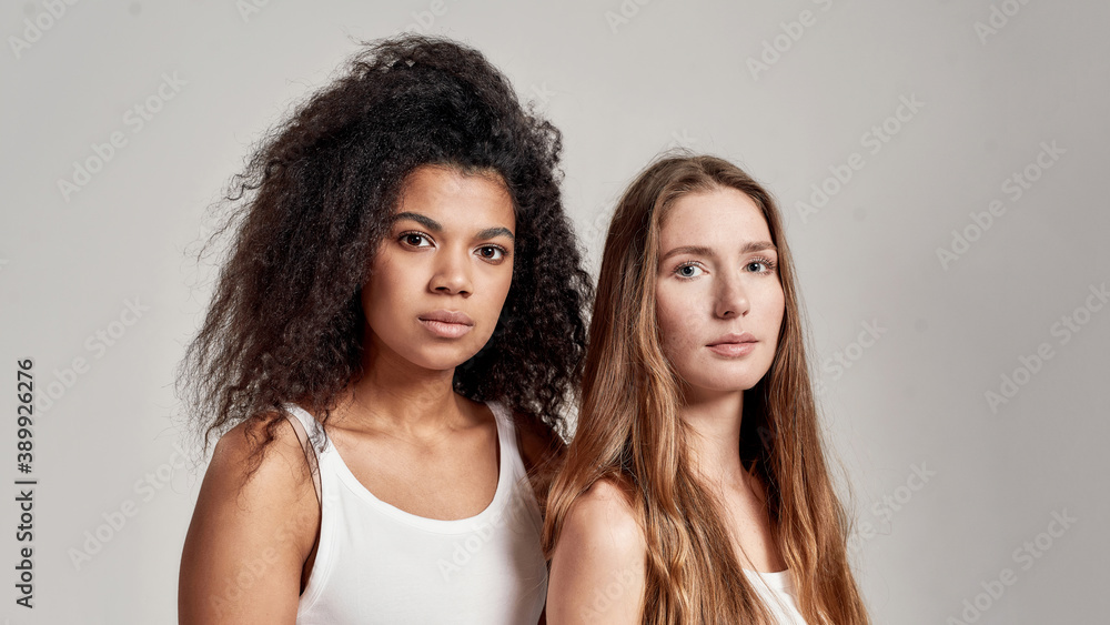 Close up portrait of two young diverse women wearing white shirts looking confident at camera while posing together isolated over grey background