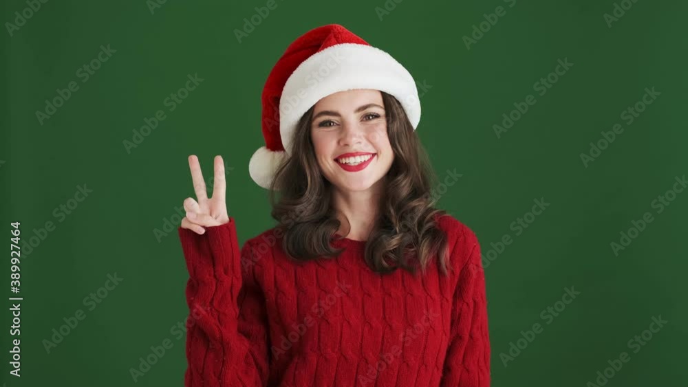 Positive girl in christmas santa hat showing peace gesture isolated over green wall background Positive girl in christmas santa hat showing peace gesture isolated over green wall background