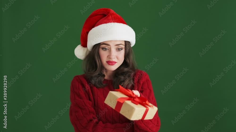 Confused stressed girl in christmas santa hat holding present box isolated over green wall background Confused stressed girl in christmas santa hat holding present box isolated over green wall background