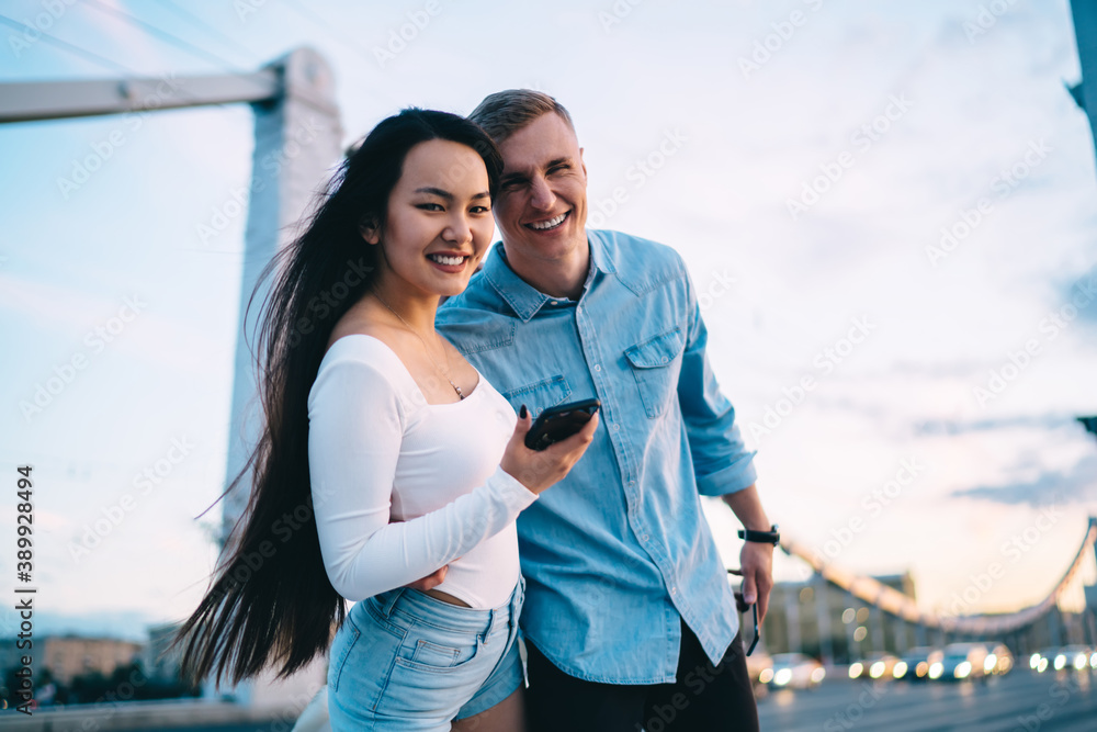 Half length portrait of cheerful couple in love smiling at camera during walking time in city, joyful hipster guys 20 years old holding smartphone device and laughing while posing at urban bridge