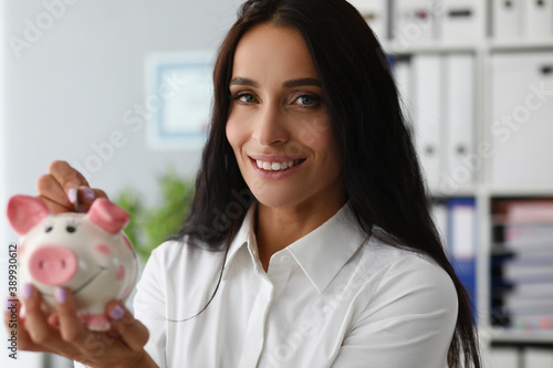 Close-up of businesslady putting money for dream in thrift-box. Smiling woman looking at camera with calmness and gladness. Piggy bank and economy concept
