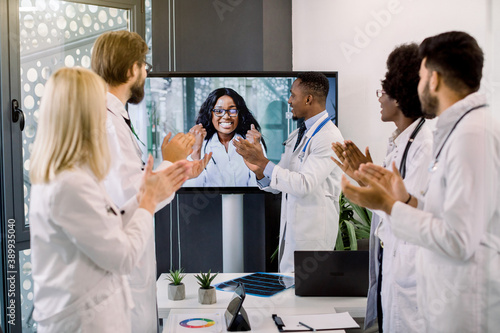 Joyful team of multiracial doctors during online video meeting indoors  clapping hands after successful presentation of their smiling excited African woman colleague  sharing results of the project