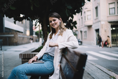 Portrait of charming hipster girl in casual clothing and electronic headphones smiling at camera while resting at urban bench and listening positive music playlist, cheerful female enjoying pastime © GalakticDreamer
