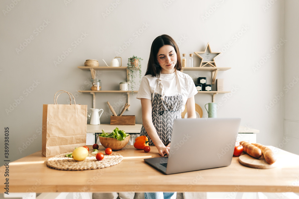 Young caucasian woman in apron use laptop computer in the modern kitchen, preparing salad, read recipe. Young caucasian woman in apron use laptop computer in the modern kitchen, preparing salad, read recipe.