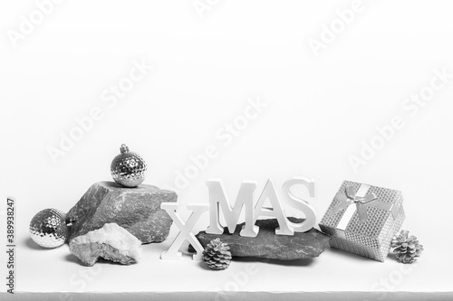 Christmas festive trendy creative minimal monochrome composition. Christmas silver and white decorations: letters XMAS, Xmas balls, pine cones, gift box on stone podiums on white. Copy space for text.