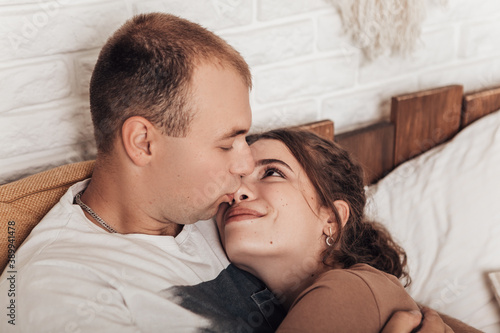 Portrait of young loving couple lying in bed, chilling and spending time together. Newlyweds kissing hugging relaxing, rest after work in cozy atmosphere indoor. Romantic concept.