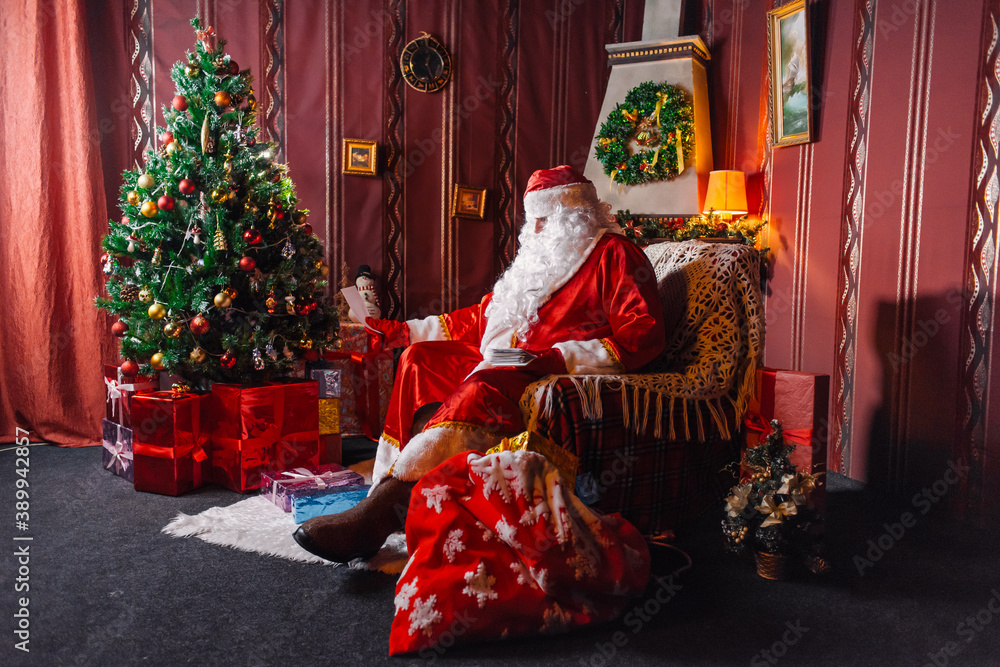 Portrait of Santa Claus - with a white beard, Santa's hat, with gifts near the Christmas tree and fireplace on an armchair on Christmas Eve and New Year. Portrait of Santa Claus - with a white beard, Santa's hat, with gifts near the Christmas tree and fireplace on an armchair on Christmas Eve and New Year.