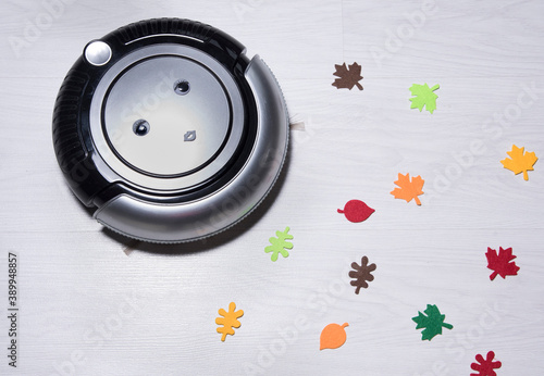Funny robot vacuum with eyes and autumn leaves on white wooden floor. Smart home concept.