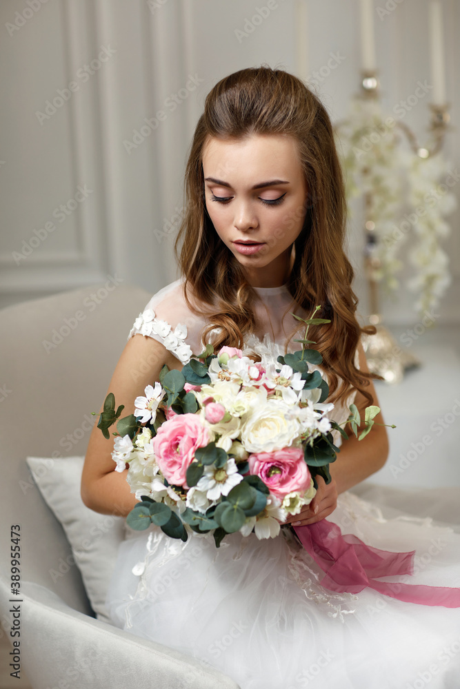 beautiful tender young bride with wedding makeup and long curly hair holding flower bouquet and sitting on armchair in white studio interior beautiful tender young bride with wedding makeup and long curly hair holding flower bouquet and sitting on armchair in white studio interior