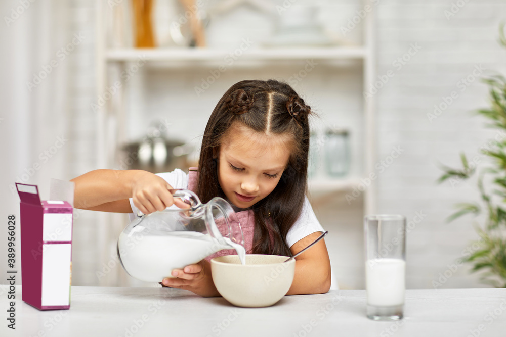 smiling cute little girl eating breakfast: cereal with the milk. child pours milk into a bowl of cereal in the kitchen. healthy breakfast smiling cute little girl eating breakfast: cereal with the milk. child pours milk into a bowl of cereal in the kitchen. healthy breakfast