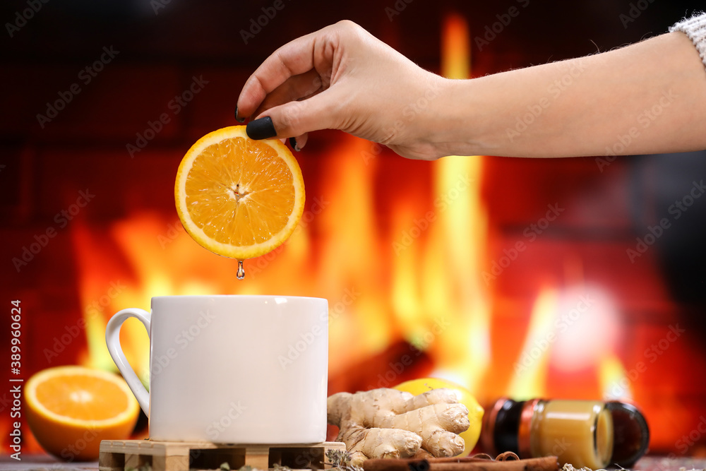 A woman's hand puts a slice of orange into white tea cup with lemon, ginger, orange and jam against the background of a burning fireplace. Healthcare. Vitamin cocktail. A woman's hand puts a slice of orange into white tea cup with lemon, ginger, orange and jam against the background of a burning fireplace. Healthcare. Vitamin cocktail.