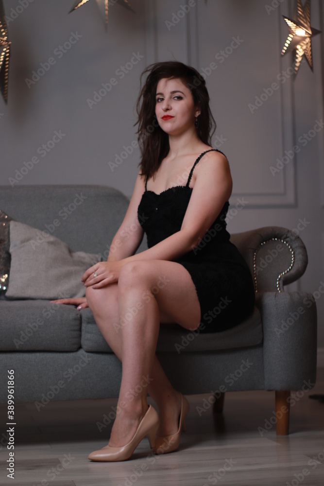 Fototapeta premium sexy attractive brunette in black skinny dress on a grey couch in a grey room with decorative big stars. studio shoot.
