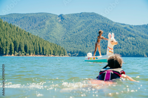 Teen girl standing on inflatable llama pool floaty on lake while sister swims with lifejacket