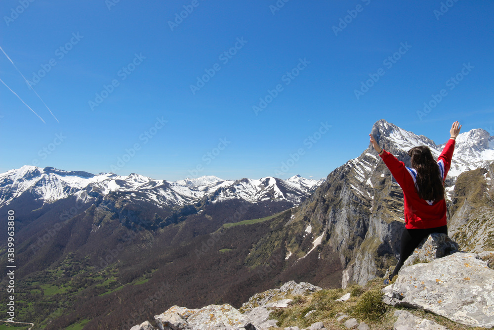 Young girl sitting on a stone with her arms raised on the edge of a cliff with mountains in the background
