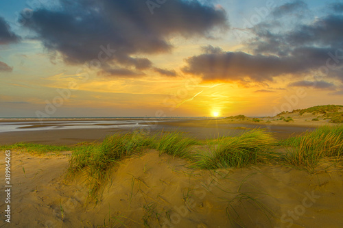 Sunset on coastline at low tide and sandy beach with wet spots and dunes with dune grass against background of blue sky with scattered clouds and sun with warm sunlight and sunbeams photo