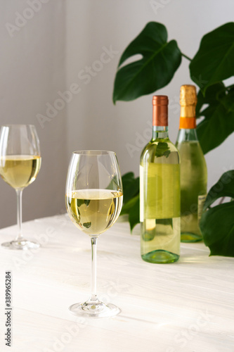 Two glasses with white wine and a bottle on a white wooden background, home flower Monstera