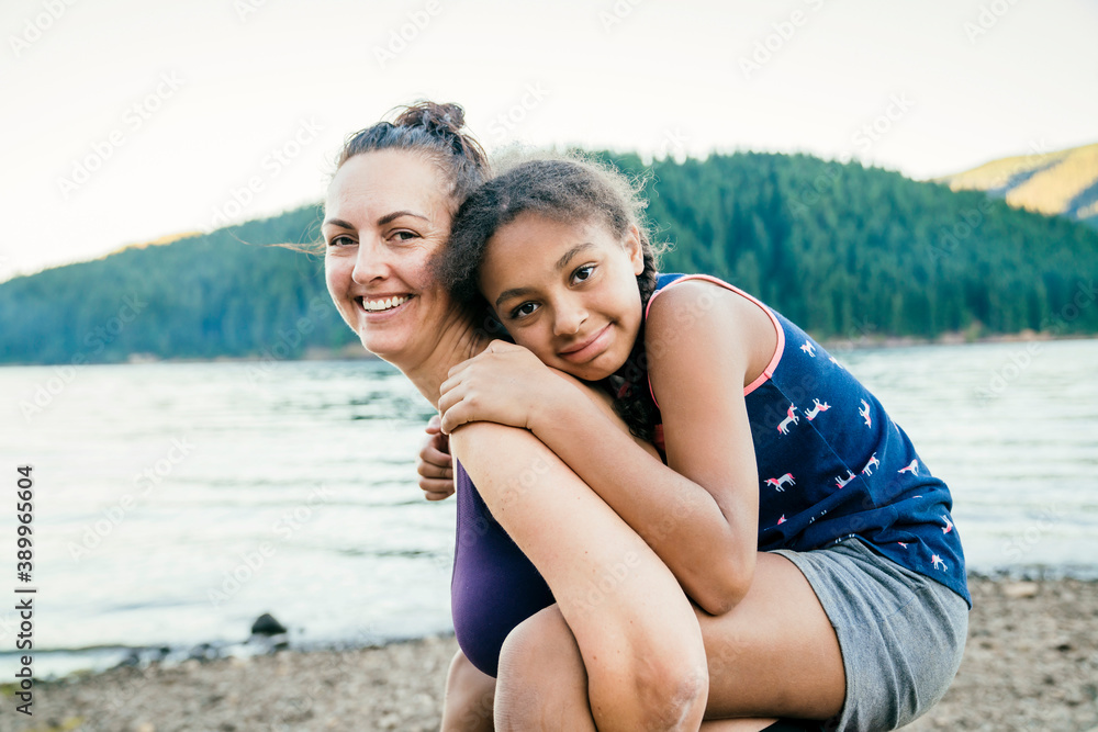 Happy mom giving daughter piggy back ride by lake in forest
